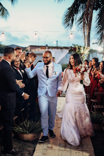 A couple dressed in wedding attire is walking hand in hand down an aisle, surrounded by smiling guests. The groom is wearing a light blue suit with a black tie, while the bride is in a white lace gown holding a bouquet of red and white flowers. String lights and palm trees are visible in the background, creating a festive atmosphere.