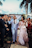 A couple dressed in wedding attire is walking hand in hand down an aisle, surrounded by smiling guests. The groom is wearing a light blue suit with a black tie, while the bride is in a white lace gown holding a bouquet of red and white flowers. String lights and palm trees are visible in the background, creating a festive atmosphere.