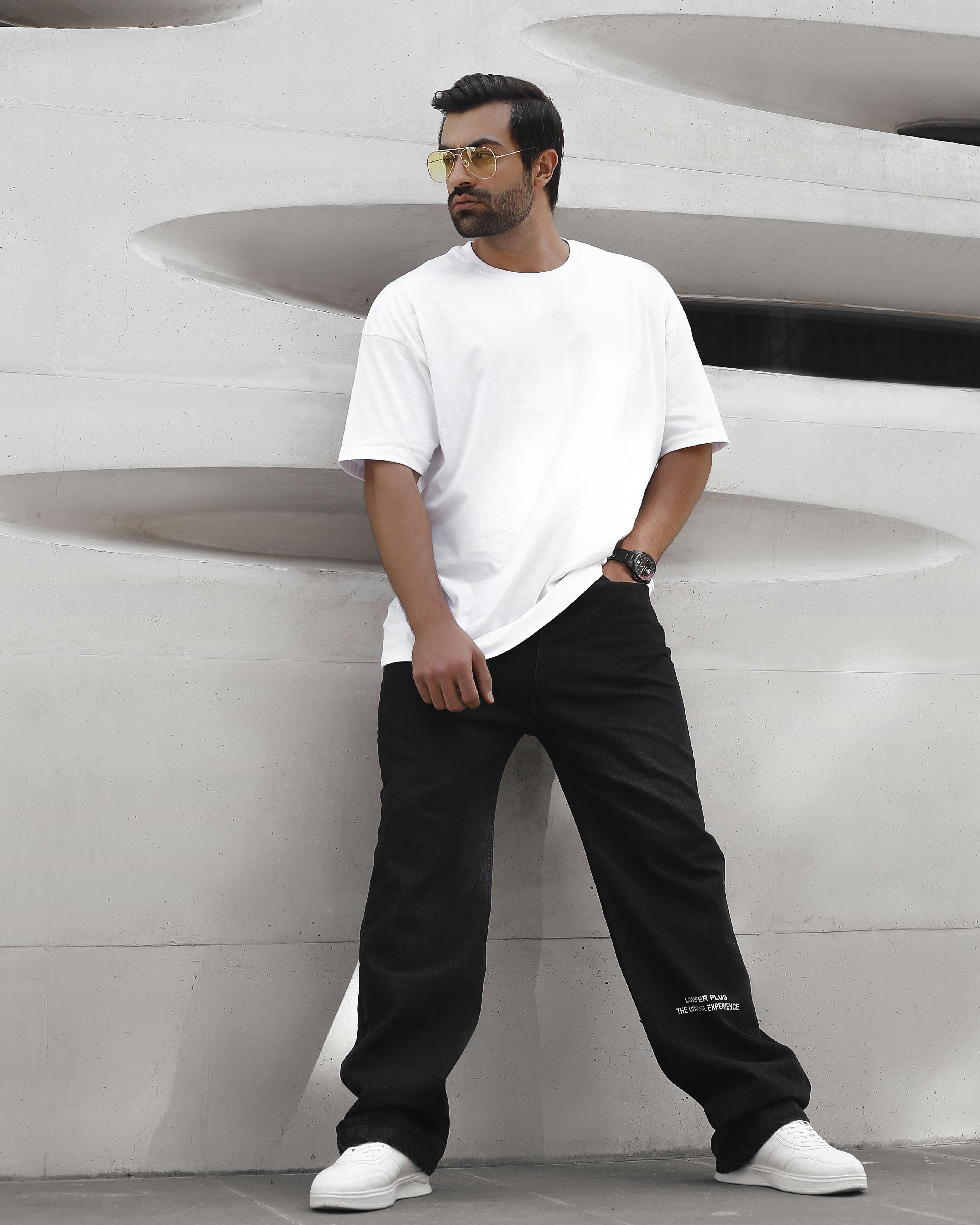 Man in relaxed white tee and wide-leg pants leaning against concrete wall, urban environment, natural light