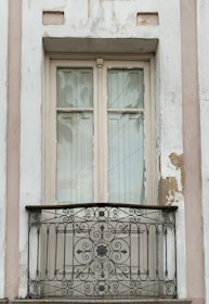 A vintage-style, cream-colored window with aged paint is surrounded by a slightly weathered wall with peeling areas. The window features two glass panels and is complemented by a decorative wrought-iron balcony railing with ornate floral and swirl designs.
