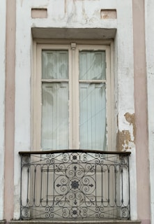 A vintage-style, cream-colored window with aged paint is surrounded by a slightly weathered wall with peeling areas. The window features two glass panels and is complemented by a decorative wrought-iron balcony railing with ornate floral and swirl designs.