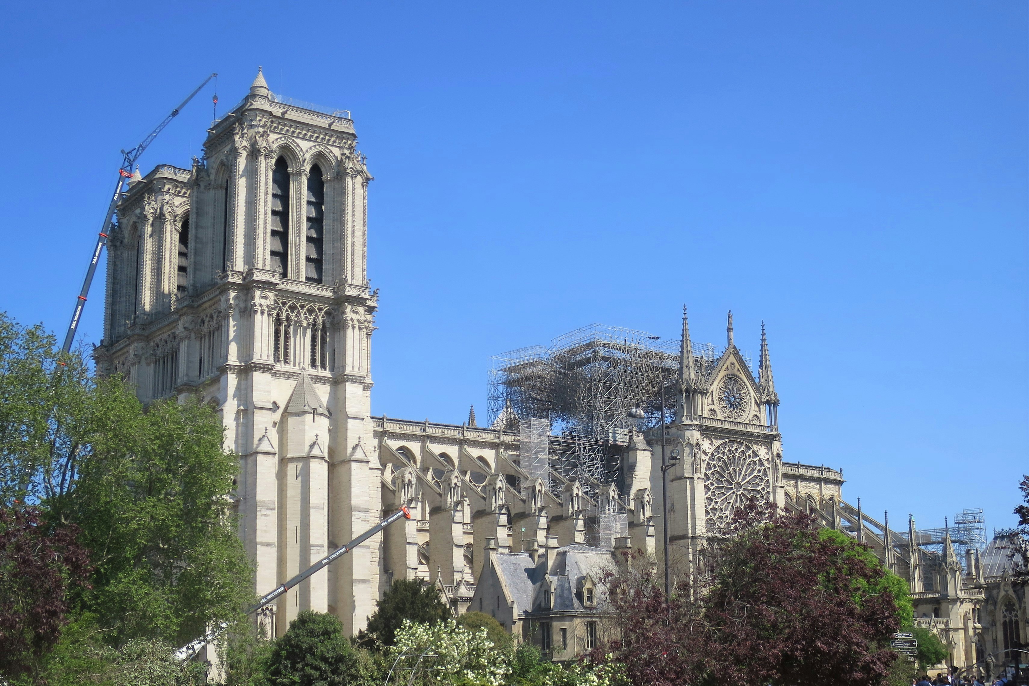 a large cathedral with a scaffolding on top of it, Notre Dame five days after the fire of 15 April 2019. The roof and spire have collapsed and the carved stone walls are charred. The remains of melted metal scaffolding remain atop the cathedral where the roof and spire were. A crane helps support a leaning bell tower. It