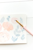 A delicate watercolor painting depicting soft pink and blue floral designs on a light background. A paintbrush with a pink handle rests on the paper, suggesting an ongoing art project.