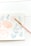 A delicate watercolor painting depicting soft pink and blue floral designs on a light background. A paintbrush with a pink handle rests on the paper, suggesting an ongoing art project.