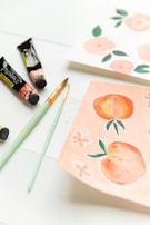 A set of art supplies including brushes and paint tubes are placed on a white surface next to two pieces of paper. One paper has watercolor illustrations of peaches and flowers, while the other features pink floral designs.