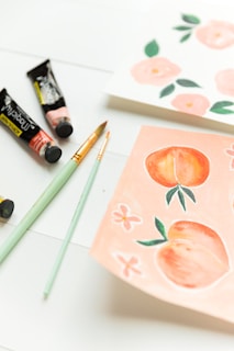 A set of art supplies including brushes and paint tubes are placed on a white surface next to two pieces of paper. One paper has watercolor illustrations of peaches and flowers, while the other features pink floral designs.