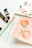 A set of art supplies including brushes and paint tubes are placed on a white surface next to two pieces of paper. One paper has watercolor illustrations of peaches and flowers, while the other features pink floral designs.