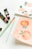A set of art supplies including brushes and paint tubes are placed on a white surface next to two pieces of paper. One paper has watercolor illustrations of peaches and flowers, while the other features pink floral designs.