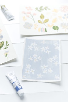 Multiple sheets of paper display watercolor botanical illustrations in pastel colors, including flowers and leaves. Tubes of paint are positioned near the sheets, suggesting an art project in progress.