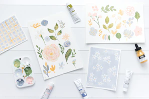 A detailed watercolor of educational printables spread out on a wooden table with pinecones and leaves nearby.