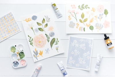 A collection of watercolor botanical illustrations on three sheets of paper, surrounded by art supplies. Pastel colors are used to depict flowers and leaves. To the left, there is a palette with various paint colors, and several tubes and a bottle of ink are scattered on a white wooden surface.