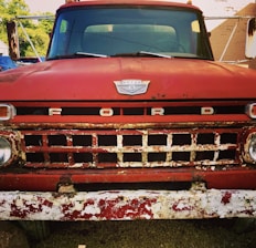 A close-up view of an old, red Ford truck's front grille, showcasing signs of rust and wear. The word 'FORD' is prominently displayed across the grille with a vintage badge above it. The truck is parked with a bit of landscape in the background.