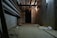 A dimly lit basement or underground passageway with exposed concrete walls and ceiling, featuring large pipes running along the left side and overhead. The floor is cluttered with various objects including an electrical outlet, plywood, and some bags. The lighting is sparse, casting shadows and creating a somewhat eerie and abandoned atmosphere.