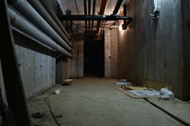 A dimly lit basement or underground passageway with exposed concrete walls and ceiling, featuring large pipes running along the left side and overhead. The floor is cluttered with various objects including an electrical outlet, plywood, and some bags. The lighting is sparse, casting shadows and creating a somewhat eerie and abandoned atmosphere.