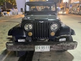 A front view of a vintage black Jeep parked on a street at night. The Jeep has a distinctive wood beam as a front bumper and a Bat emblem on the grille. The license plate reads '9511'. Surrounding the vehicle are urban elements, such as parked motorcycles, street lights, and buildings.