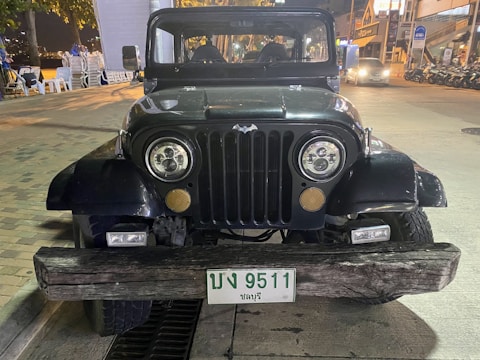 A front view of a vintage black Jeep parked on a street at night. The Jeep has a distinctive wood beam as a front bumper and a Bat emblem on the grille. The license plate reads '9511'. Surrounding the vehicle are urban elements, such as parked motorcycles, street lights, and buildings.