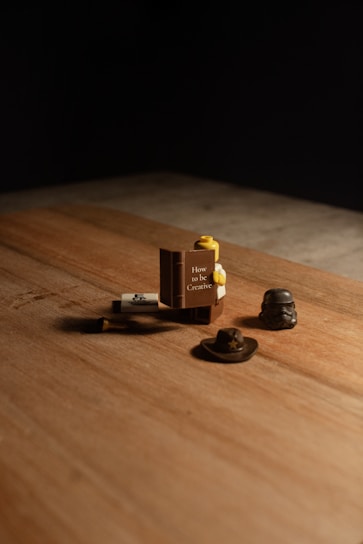 A wooden surface with a set of small objects, including a Lego piece shaped like a book titled 'How to be Creative'. Nearby are a small hat and a helmet resembling a stormtrooper helmet.