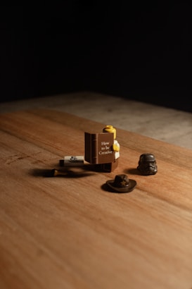 A wooden surface with a set of small objects, including a Lego piece shaped like a book titled 'How to be Creative'. Nearby are a small hat and a helmet resembling a stormtrooper helmet.