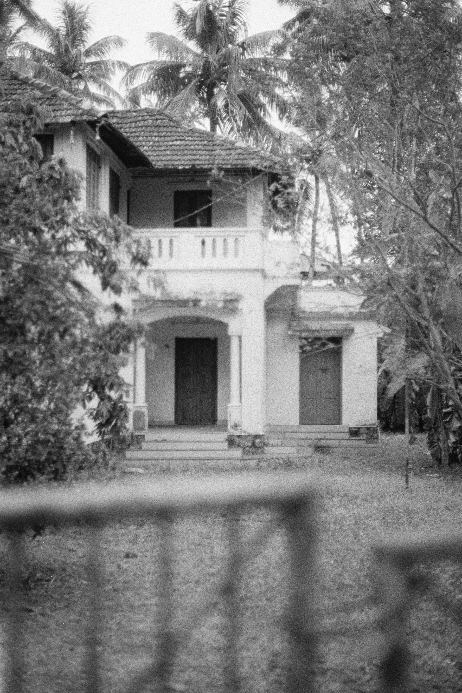 A warm, nostalgic sepia-toned photograph of Annamma's old family home surrounded by lush greenery, evoking memories of simpler times.