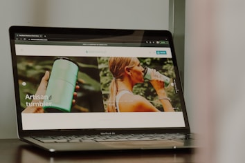 A photograph of a laptop displaying a website that features a product named 'artisan tumbler'. The website has images of a person holding a green tumbler and another image of a person drinking from the tumbler. The laptop brand is MacBook Air.