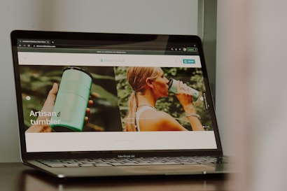 A photograph of a laptop displaying a website that features a product named 'artisan tumbler'. The website has images of a person holding a green tumbler and another image of a person drinking from the tumbler. The laptop brand is MacBook Air.