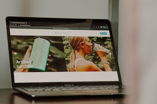A photograph of a laptop displaying a website that features a product named 'artisan tumbler'. The website has images of a person holding a green tumbler and another image of a person drinking from the tumbler. The laptop brand is MacBook Air.
