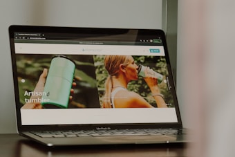 A photograph of a laptop displaying a website that features a product named 'artisan tumbler'. The website has images of a person holding a green tumbler and another image of a person drinking from the tumbler. The laptop brand is MacBook Air.