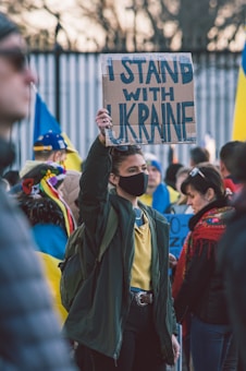 A person wearing a black mask holds a sign that reads 'I STAND WITH UKRAINE' among a group of people, some of whom are holding or wearing items in blue and yellow, the colors of the Ukrainian flag.