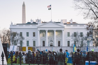 A large building with classical architecture, featuring tall white columns, is set against a clear sky. In the foreground, a diverse group of people are gathered, holding signs and flags, including those in blue and yellow. The setting appears to be a formal and significant location. A monument and an American flag are visible, adding to the atmosphere of national importance.