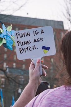 A person holds up a sign with a handwritten message expressing a strong negative opinion about Putin. The message includes a small heart with the colors blue and yellow, possibly representing the Ukrainian flag. The background is an urban setting with blurred buildings.