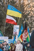 A group of people participating in a protest or rally, holding various flags and signs. The flags include those of Ukraine and Poland, suggesting a show of international solidarity. There are several prominently displayed signs with messages of support and protest.