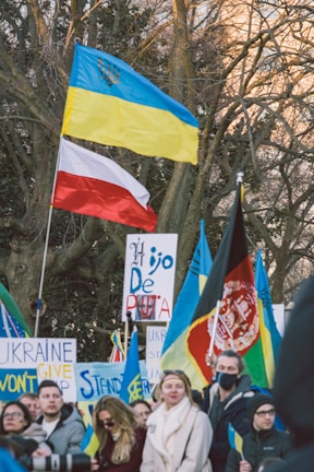 A group of people participating in a protest or rally, holding various flags and signs. The flags include those of Ukraine and Poland, suggesting a show of international solidarity. There are several prominently displayed signs with messages of support and protest.