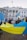 A group of people holding a large blue and yellow flag stand in front of a white building with columns and multiple windows. One participant holds a sign reading 'Military Support for Ukraine'. An American flag flies above the building.