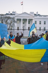 A group of people holding a large blue and yellow flag stand in front of a white building with columns and multiple windows. One participant holds a sign reading 'Military Support for Ukraine'. An American flag flies above the building.