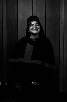 A person is wearing graduation attire, including a cap and gown, standing in front of a wooden panel wall. They are smiling broadly and wearing glasses.