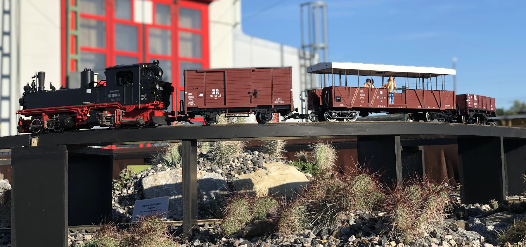 A black model steam engine pulling three red-brown cars across a low dark bridge above rocks and tufts of ornamental grass