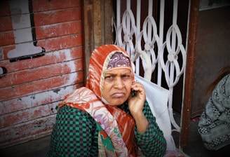 An elderly woman with a serious expression sits against a textured, worn wall. She is wearing a red-orange scarf around her head and a green checkered garment. One hand is raised towards her ear, possibly holding a device. The setting appears rustic with a metal grill and wooden elements visible in the background.