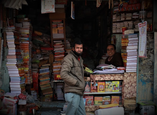 A small bookshop filled with numerous stacks of books, including large textbooks and colorful magazines. Two men are present; one is standing outside wearing a jacket, and the other is sitting inside at a desk, smiling. Calendars and posters are visible on the walls, and the shelves are overflowing with a variety of books.