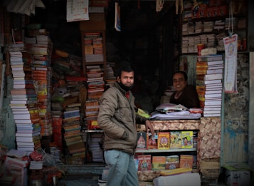 A small bookshop filled with numerous stacks of books, including large textbooks and colorful magazines. Two men are present; one is standing outside wearing a jacket, and the other is sitting inside at a desk, smiling. Calendars and posters are visible on the walls, and the shelves are overflowing with a variety of books.