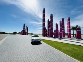 A modern outdoor installation features an array of tall, rectangular pink and white pillars arranged in a linear pattern. A white car is parked on a paved road next to a patch of neatly maintained green grass. The sky is clear with a few wispy clouds, and palm trees are visible in the background.