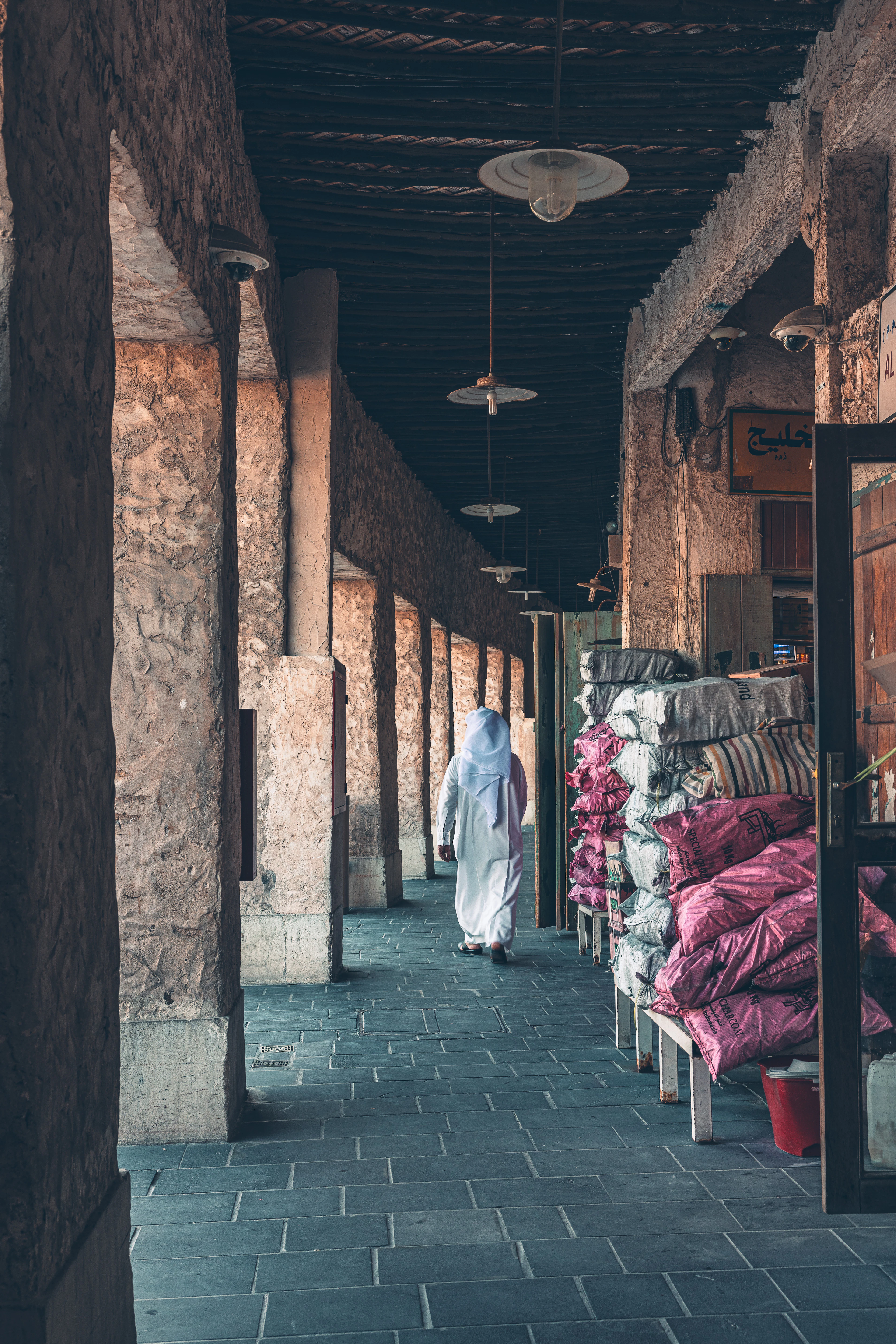 A man in a white thobe walking away down a shaded stone colonnade in Souq Waqif, Doha, past stacks of magenta and pink bolster cushions piled beside the pillars