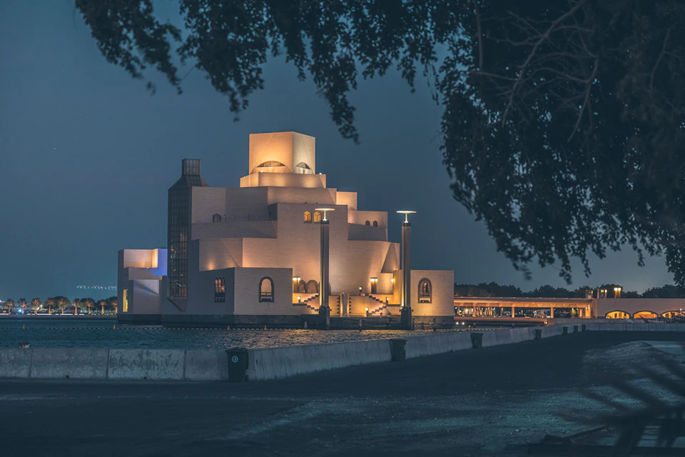 The image depicts a large, white building with multiple levels and windows, standing out against the dark sky at night. The building is illuminated by warm yellow lights that cast a soft glow on its exterior. A small tree can be seen in front of the building, adding a touch of nature to the urban landscape. In the background, there are other buildings visible, suggesting that this structure might be part of a larger complex or cityscape.