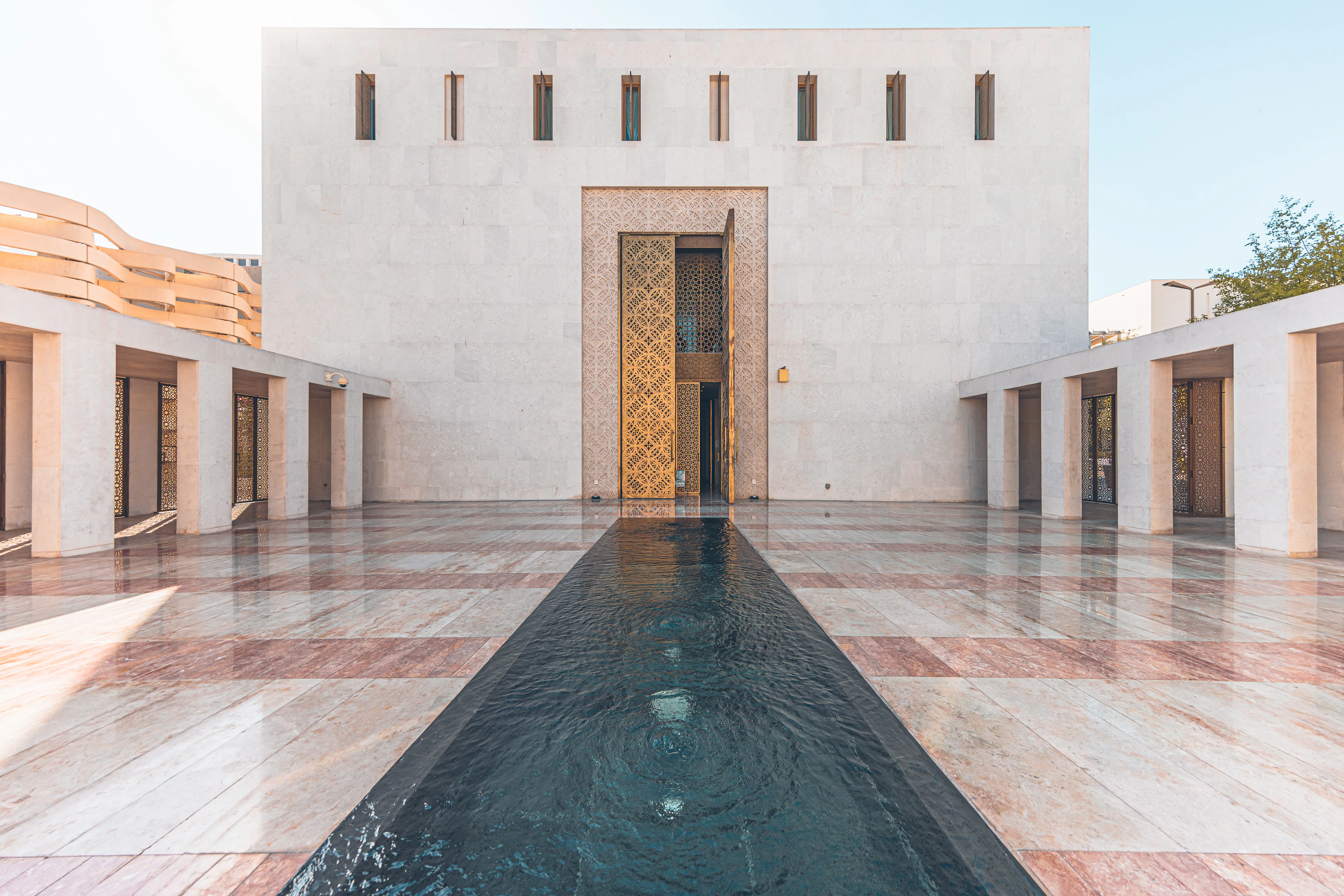 A long still reflecting channel running across a chequered marble courtyard in Msheireb Downtown Doha toward a plain white stone facade pierced by a tall gilded lattice door, with the curved sand-coloured discs of the National Museum just visible beyond