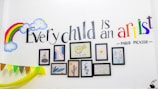 A wall decorated with a colorful rainbow and smiling clouds. The quote 'Every child is an artist - Pablo Picasso' is elegantly painted, with 'artist' in vibrant multicolors. Below the text, several framed art pieces including abstract designs and a drawing of an owl are arranged neatly. Bunting with multicolor flags is draped along the wall.
