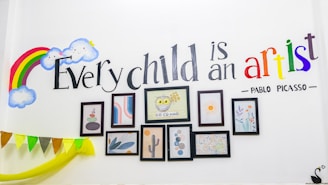 A wall decorated with a colorful rainbow and smiling clouds. The quote 'Every child is an artist - Pablo Picasso' is elegantly painted, with 'artist' in vibrant multicolors. Below the text, several framed art pieces including abstract designs and a drawing of an owl are arranged neatly. Bunting with multicolor flags is draped along the wall.