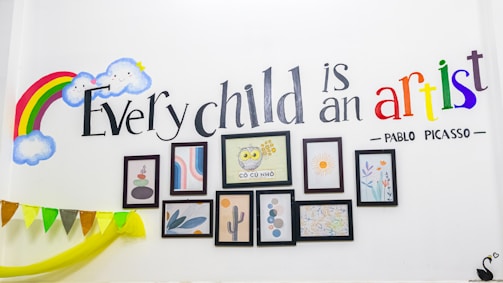A wall decorated with a colorful rainbow and smiling clouds. The quote 'Every child is an artist - Pablo Picasso' is elegantly painted, with 'artist' in vibrant multicolors. Below the text, several framed art pieces including abstract designs and a drawing of an owl are arranged neatly. Bunting with multicolor flags is draped along the wall.