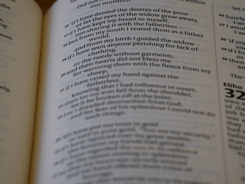 A close-up view of an open book page with text that appears to be from a religious scripture or a literary work. The text is printed in black on a white background with some parts emphasized in a bolder font. The background is blurred, focusing on the central portion of the text.