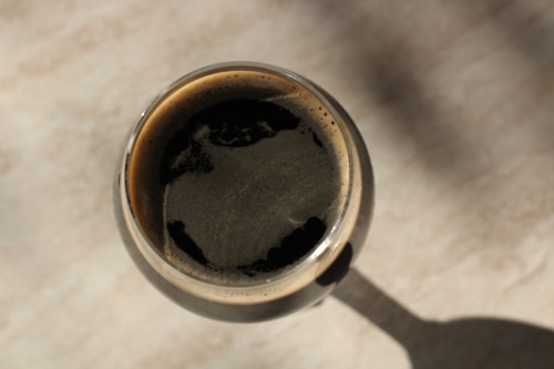 A top-down view of a glass of dark stout beer, with a frothy head and a smooth, dark surface. The shadow of the glass extends on a light-colored surface.
