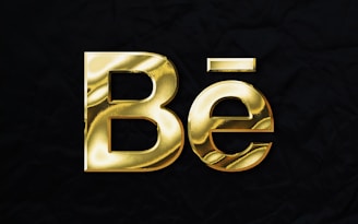 Gold metallic text displaying 'Bē' against a black textured background.