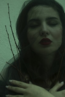 A person with closed eyes, wearing dark makeup and red lipstick, is partially wrapped in a sheer black fabric. Sparse, thin branches protrude around the face, adding an element of nature. The greenish lighting gives a serene yet mysterious feel, with the individual's hands crossed over their chest.